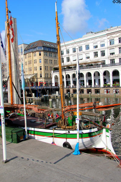Green & white canal boat docked on Kleiner Alster canal before Venetian-style arcade on opposite shore. Hamburg, Germany.